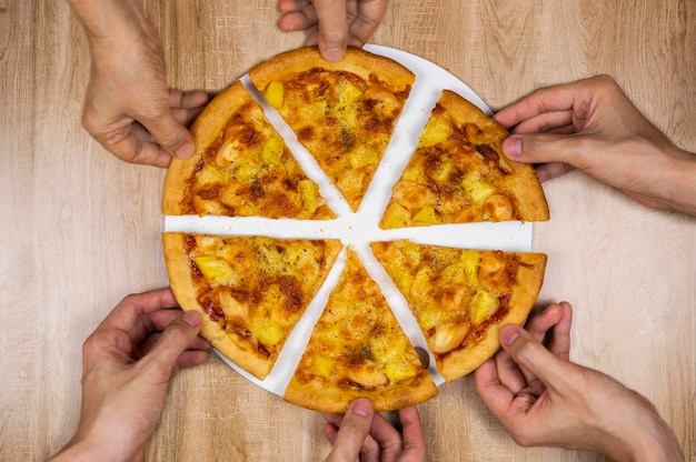 Top view of hands taking slices of delicious and crispy hawaiian pizza Group of hungry friends sharing delicious lunch on wooden table