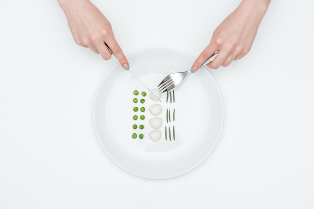 Top view of hands of young woman eating green peas, mozzarella and herbs using fork and knife