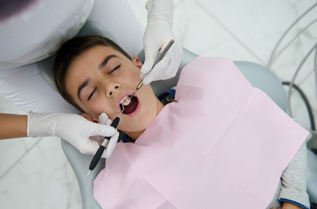 Photo top view of handsome boy at a dental check-up in white dental clinic with modern equipment. pediatric dentist examining child's teeth using sterile dental instruments - mirror and periodontal probe