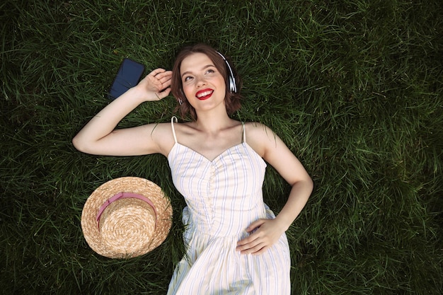 Top view of happy woman in dress and headphones lying on grass while listening music and looking away outdoors