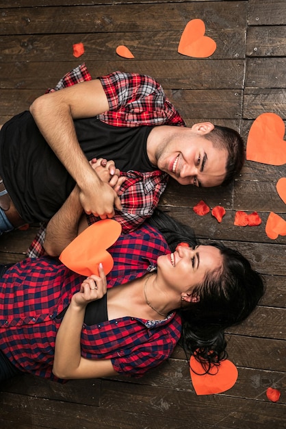 Top view of happy young couple looking at each other and smiling while lying on wooden floor Girl is holding a red paper heart