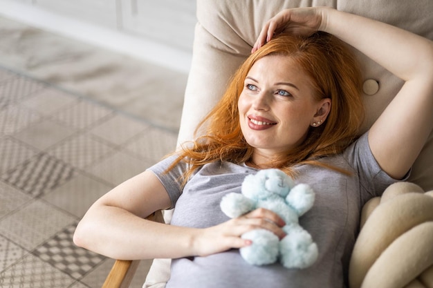 charming redhead pregnant woman with headphones on her belly sitting on  sofa with teddy bear, Stock image