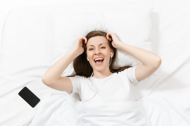 Top view of happy young woman lying in bed with white sheet, pillow, blanket, listen music from earphones in mobile phone