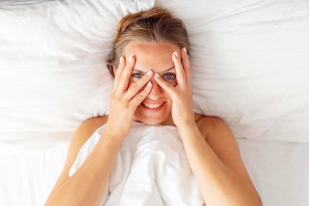 Top view of happy young woman peeking through her fingers and smiling while lying in bed