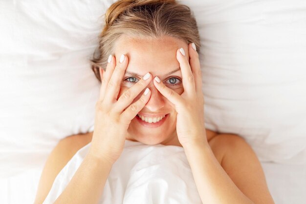 Top view of happy young woman peeking through her fingers and smiling while lying in bed