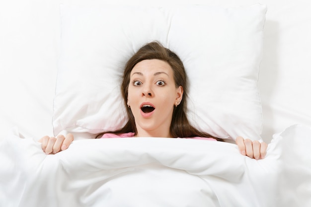 Top view of head of happy brunette young woman lying in bed with white sheet, pillow, blanket. Fun pretty female spending time in room