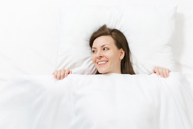 Top view of head of happy brunette young woman lying in bed with white sheet, pillow, blanket