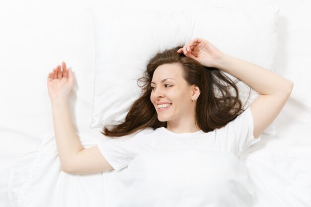 Top view of head of happy brunette young woman lying in bed with white sheet, pillow, blanket