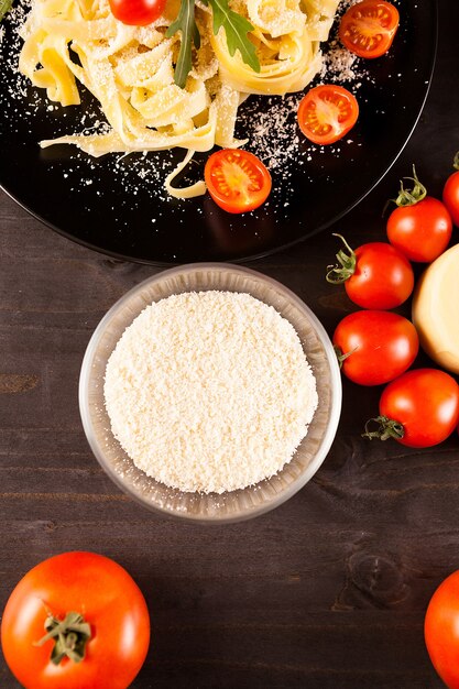 Top view of healthy and delicious tagliatelle in black plate next to greenery, cherry tomatoes and parmesane on wooden background