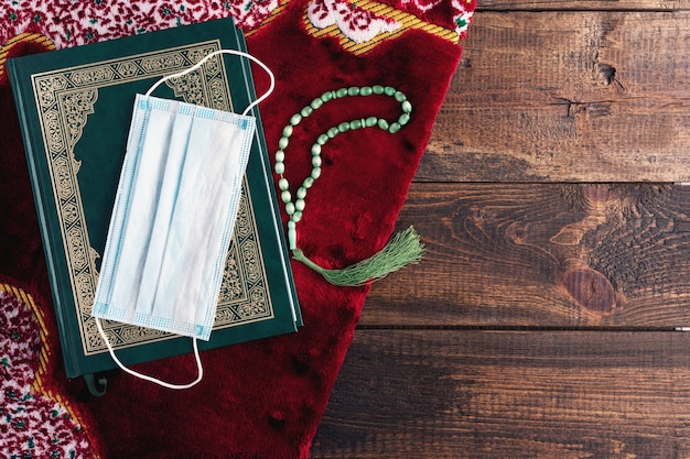 Top view Holy book Koran, rosary, medical mask on red carpet on brown wooden background, Ramadan concept, Holy month under quarantine, copy space