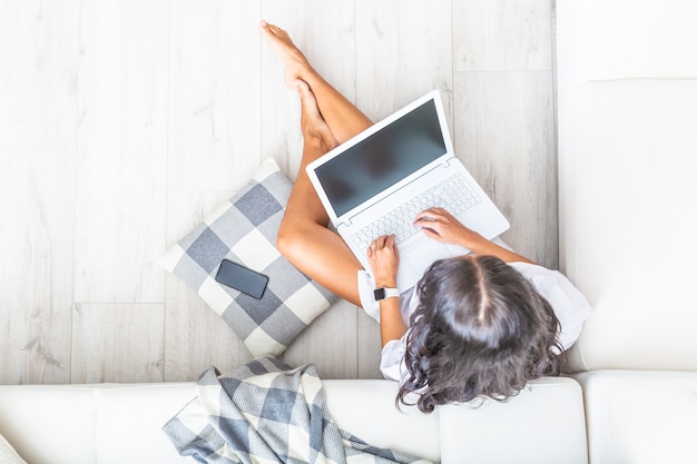 Top view of home office woman, working on a notebook, leaned against a white sofa, looking into the pc, bare legs, her cell phone next to her on a grey and white checked pillow.