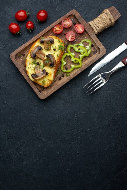 top view of homemade tasty snack with mushrooms and chopped vegetables on wooden board cutlery set on black background