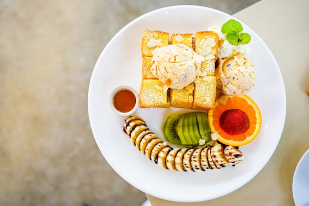 top view of honey toast bread served with mixed fruits, sliced banana, ice-cream and topped with almond slice and honey syrup in white plate.