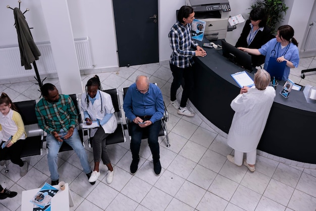 Top view of hospital waiting area with senior doctor admitting asian patient into clinic at front desk. African american doctor filling form for patient appointment in medical healthcare facility.