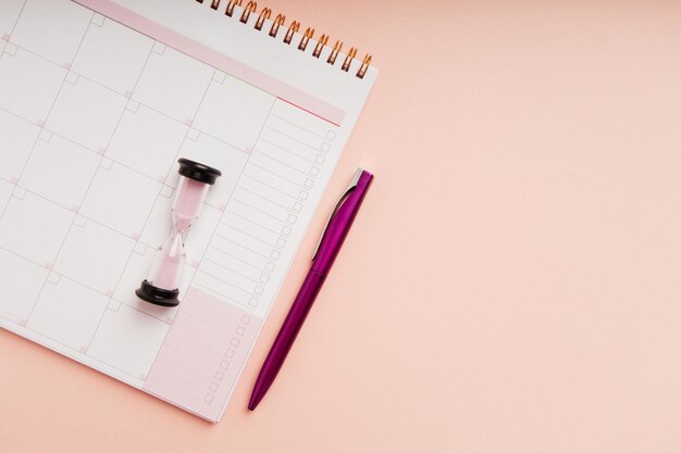 Top view of hourglass with month planner and pen on a pink background Time management and schedule