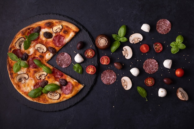 Vista dall'alto della pizza affamata che mangia mozzarella, funghi, salsicce, basilico, olive, pomodori. il concetto piatto di pizza mangia ingredienti su sfondo di cemento nero