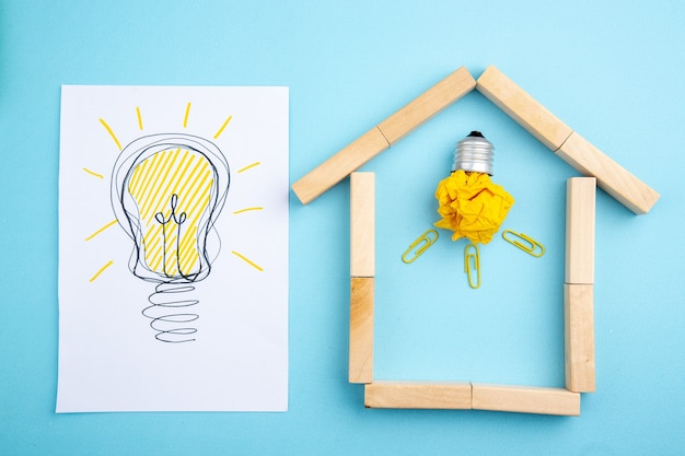 写真 青い背景の上の家の形をした木製ブロックのアイデア電球の概念と紙のしわくちゃの紙に描く上面図idealight電球