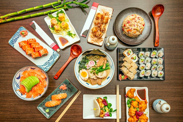 Top view image of Asian dishes featuring salmon sashimi with avocado fish and vegetable ramen assorted trays of uramaki and maki California roll soy sauce bao bread