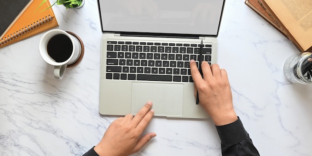 Photo top view image of businessman's hands using a computer tablet with white blank screen that putting on marble texture table with coffee cup, notebook, books, potted plant and pencil holder.