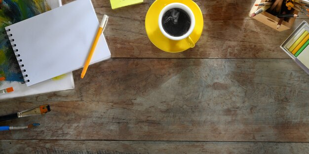 The top view image of a coffee cup is putting on a wooden table surrounded by painting equipment.