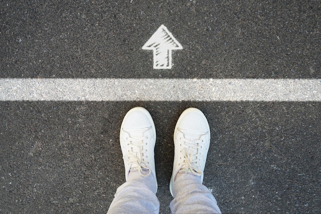 Top view image of female legs in white sneakers and white arrow on asphalt road