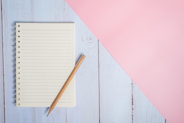 Top view image of notebook with pencil on wooden table with pink background