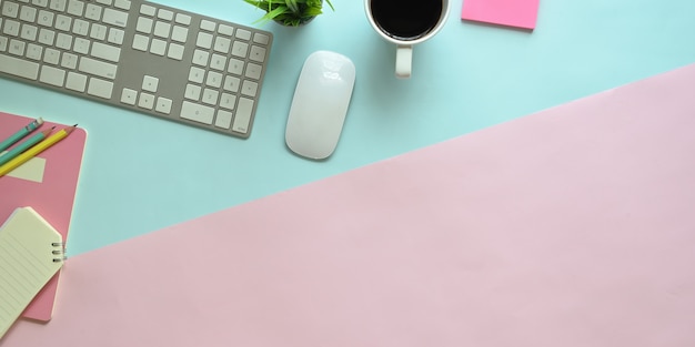 Top view image of pastel color working table with office equipment putting on it. Flat lay keyboard, wireless mouse, coffee cup, notebook, potted plant and pencils. Adorable workplace concept.