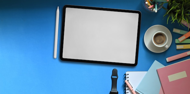 Top view image of white blank screen tablet putting on colorful working desk that surrounded by marker pens, notebooks, smartwatch, coffee cup, pencil holder and potted plant. Cluttered workspace.