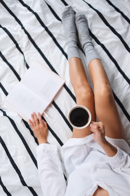 Top view image of a woman reading book and drinking hot coffee in a white cozy bed at home in the morning