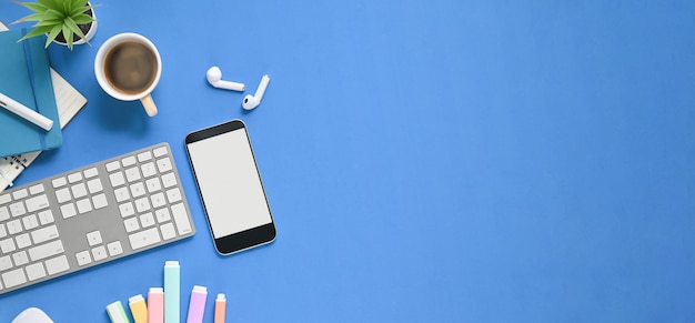 Top view image of working table with accessories putting on it. White blank screen smartphone, coffee cup, wireless earphone, notebook, pen and wireless keyboard flat lay on blue space.