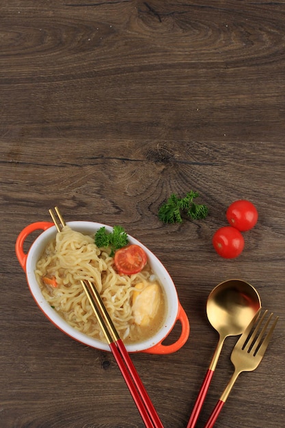 Top View Indomie, 젓가락으로 오렌지 그릇에 계란, 토마토, 다진 고추를 곁들인 맛있는 인도네시아 인스턴트 국수. 복사 공간이 있는 테이블에서