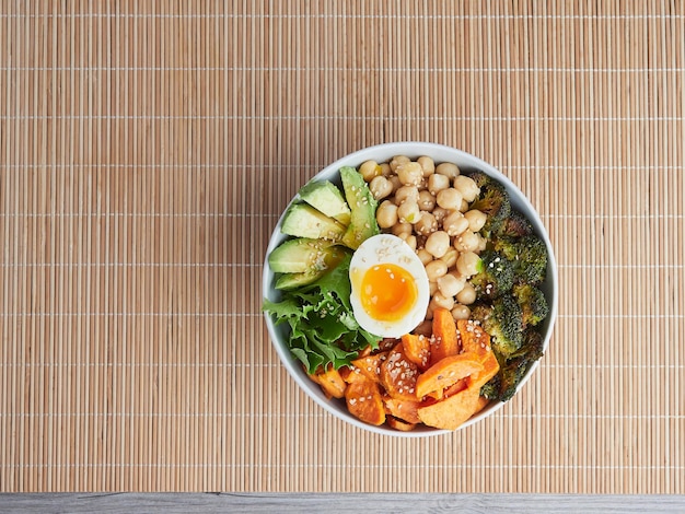 Top view of an isolated Buddha Bowl on bamboo tablecloth with chickpeas, avocado, roasted broccoli and sweet potato, lettuce and boiled egg, sesame seeds on top. Healthy vegetarian meal. Copy space