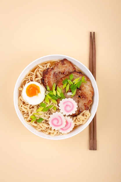 Top view of Japanese noodle soup ramen in white ceramic bowl with noodles, meat broth, sliced roasted pork, green onion, narutomaki, egg yolk, beige background. Traditional dish of Japan, close-up