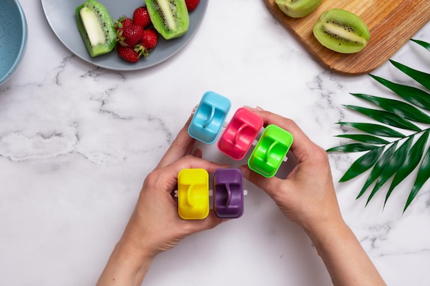 Vista dall'alto del tavolo da cucina con stampi per gelato ripieni pronti per il congelamento in mani femminili produzione di gelato fatto in casa con yogurt, fragole e kiwi