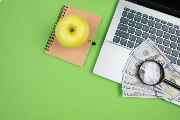 Photo top view of laptop dollars magnifying glass notepad and apple on green background