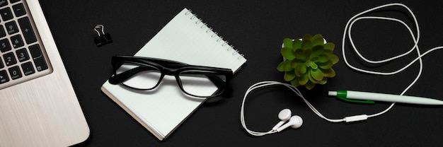 Top view of the laptop, glasses, notebook, headphones and a green plant on a black background. The concept of business, freelancing, remote work or work in the office. banner.