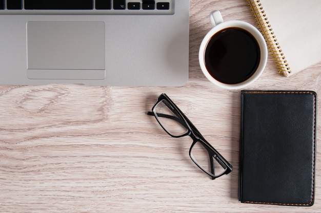 Vista superiore del computer portatile e del blocco note con i vetri e il libro tascabile sul desktop e sulla tazza di caffè di legno.