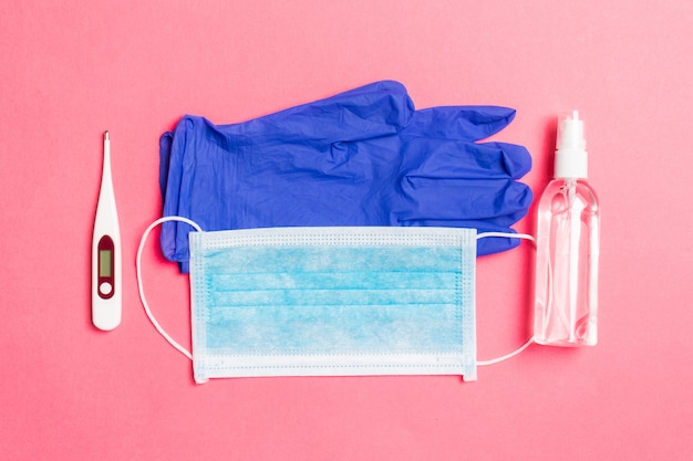 Top view of latex gloves, bottle of alcohol hand sanitizes, medical mask and digital thermometer on pink