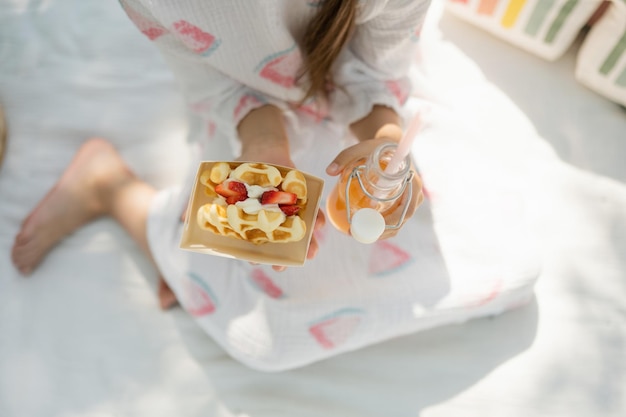 Top view of a little girl in a dress sitting on a blanket and holding strawberry waffles and juice