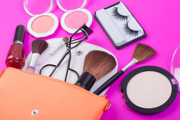 Top view of a make up bag, with cosmetic beauty products spilling out onto a  pink background.