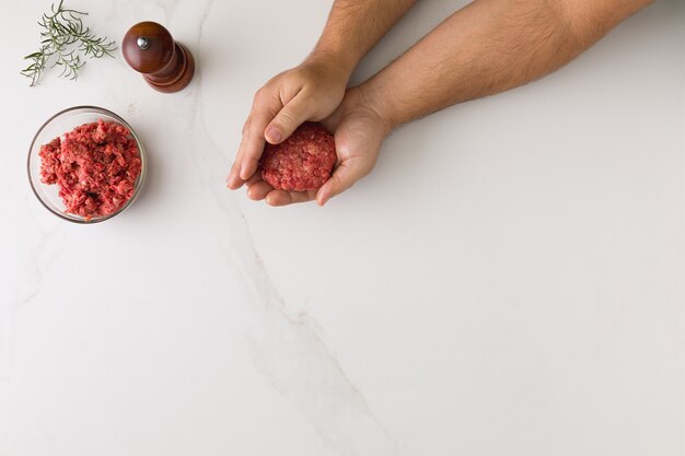 Top view of male hand molding a hamburger in a marble table, a glass bowl with meat, wooden pepper grinder and rosemary with space for text