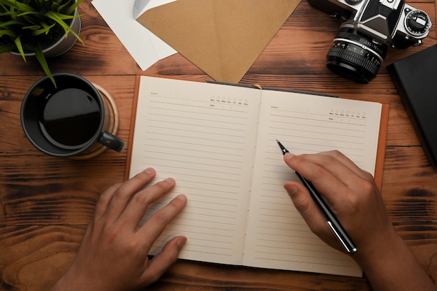 Top view of male hand writing on blank notebook on wooden table with camera and coffee cup