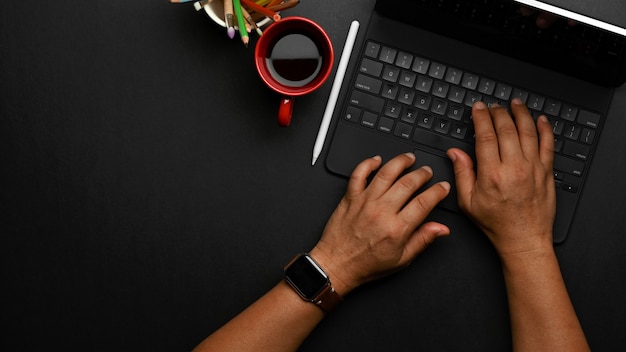 コーヒーカップと黒いテーブルの上のタブレットキーボードで入力する男性の手の上面図