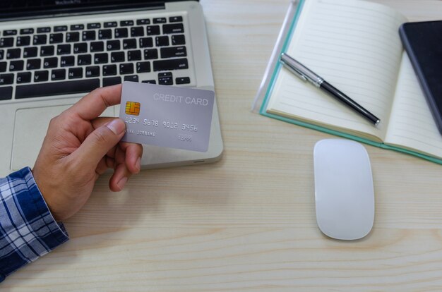 Top view man holding a credit card and wearing a blue plaid shirt on the desk. Laptop computer Notepad and pen.Online shopping business, pay by credit card