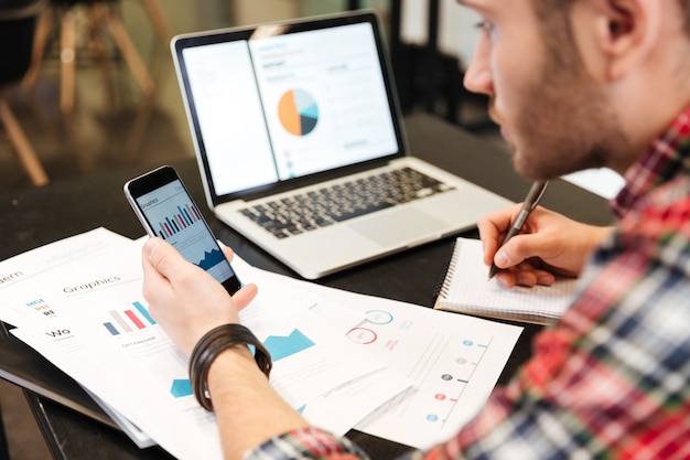 Top view of man sitting at the table, analyzes performance and looking at phone in office.