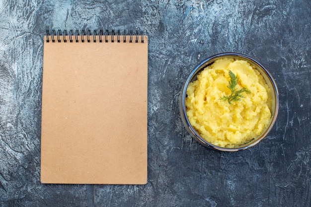 Top view of mashed potatoes served with dill and notebook on dark color background with free space