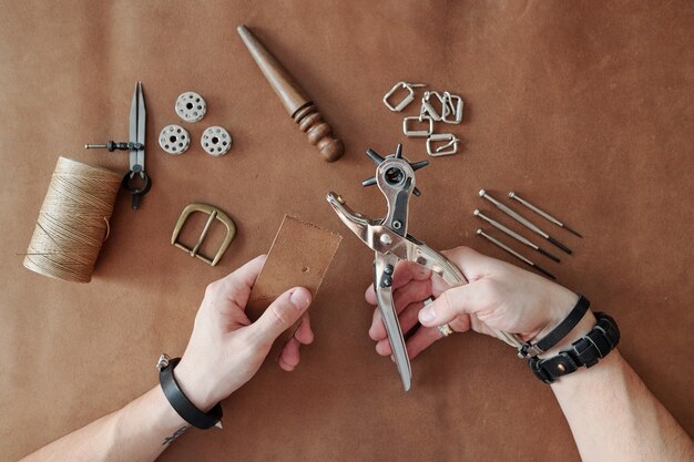 Top view of master hands in bracelets holding handtool for making holes in leather or suede during working process