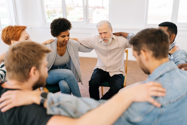 Top view of mature male therapist and group of multiage patients putting hands on each others shoulders sitting in circle during group therapy session