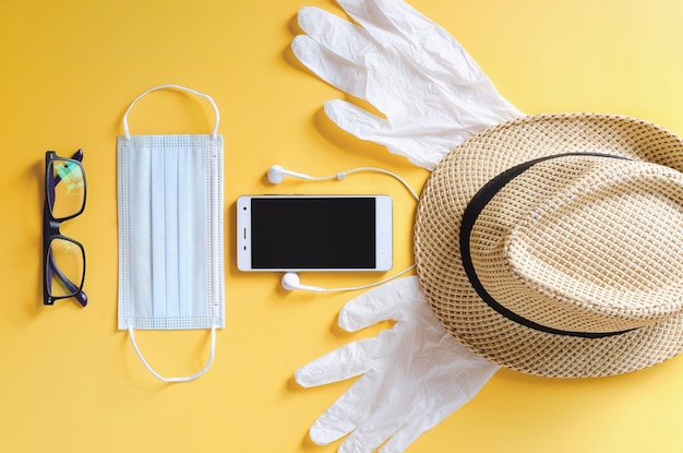 Top view of medical face mask, gloves, phone, beach hat. yellow background. Travel after Covid-19.