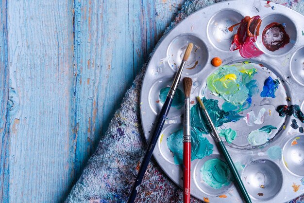 Top view of a metal painting pallet with brushes on a cloth and weathered blue wooden surface